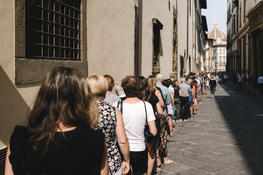 A long queue of visitors stretching down a narrow Florentine street beside a plain museum facade on a bright morning