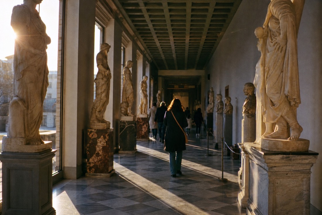Classical marble sculptures lining a sunlit gallery hall in Florence with a coffered ceiling overhead