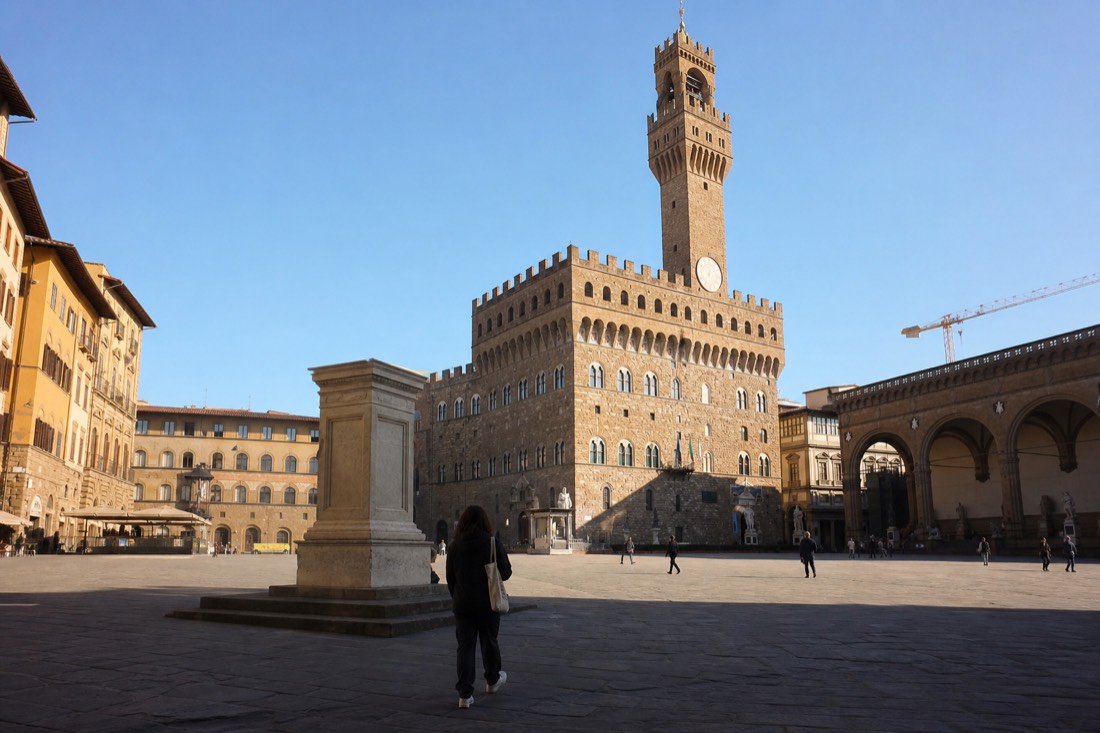 A wide cobbled Florentine square at dawn with the crenellated tower of Palazzo Vecchio rising behind an empty stone pedestal