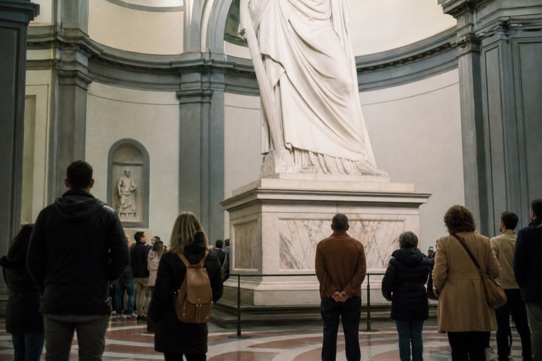 Visitors standing at floor level beside the tall stone pedestal of David in the Accademia Tribuna, their heads reaching only to the base of the statue