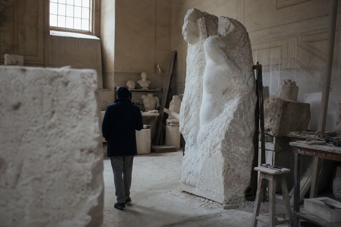 Dim sculpture studio with an unfinished marble figure emerging from rough stone beneath a high window