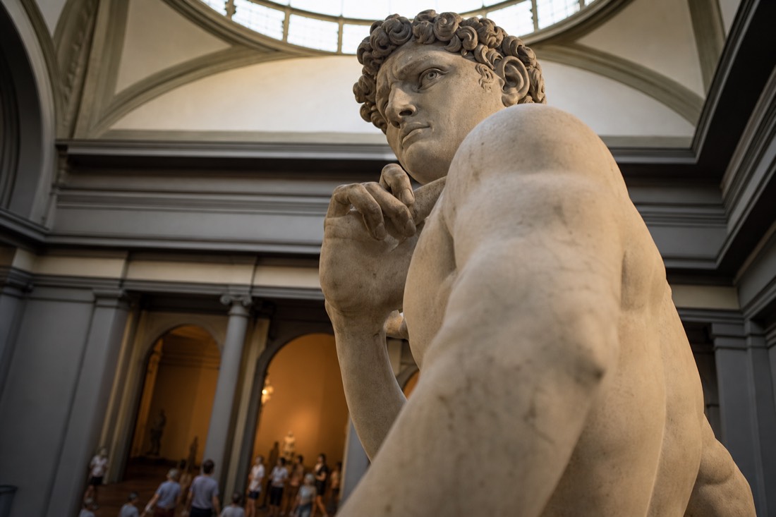 Detail of a tall marble statue's tense right hand and furrowed brow under bright museum skylight in Florence