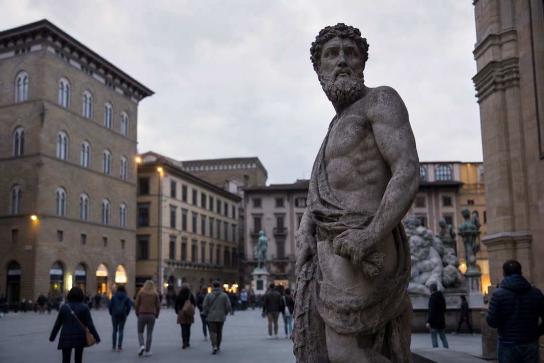 A weathered stone statue standing exposed in an open Florentine piazza under a bright overcast sky with old facades behind