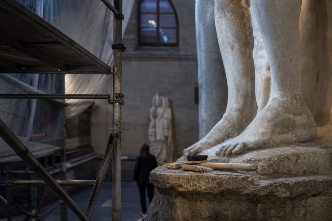 A conservator's scaffold and soft brushes beside the lower legs of a tall marble statue in a museum conservation setting