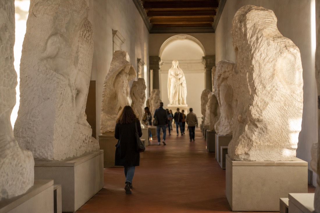 The Hall of the Prisoners in the Accademia Gallery with unfinished marble figures lining a corridor that frames the distant statue of David