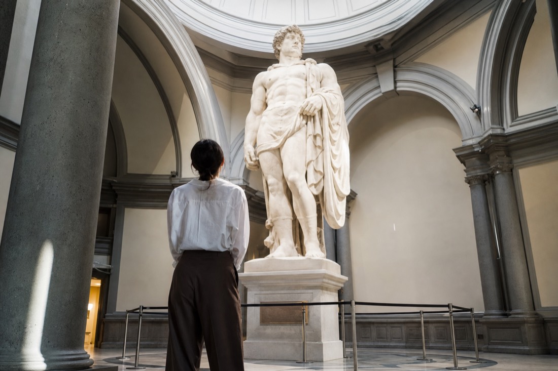 A single visitor standing beneath the towering marble statue of David in the skylit tribune of the Accademia Gallery in Florence