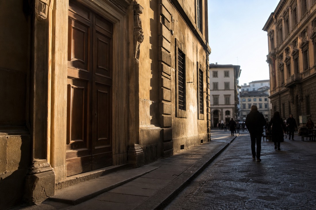 Closed wooden door of a Florentine museum entrance on a quiet cobbled street with morning shadows