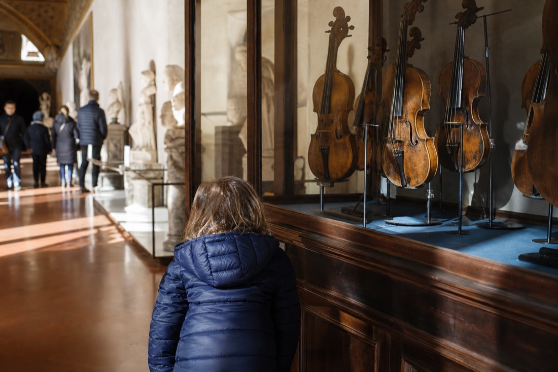 A child leaning toward a glass case of antique violins in the musical instruments room of the Accademia Gallery in Florence