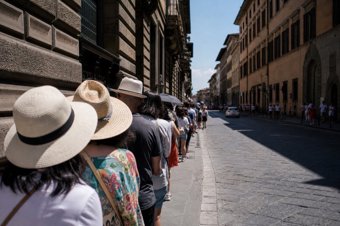 Queue of visitors with sunhats waiting along the shaded side of Via Ricasoli in Florence at midday