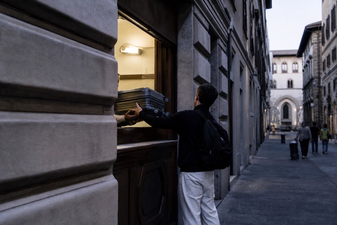 A traveler handing a suitcase across the counter of a small luggage-storage office on a Florence street near the Accademia