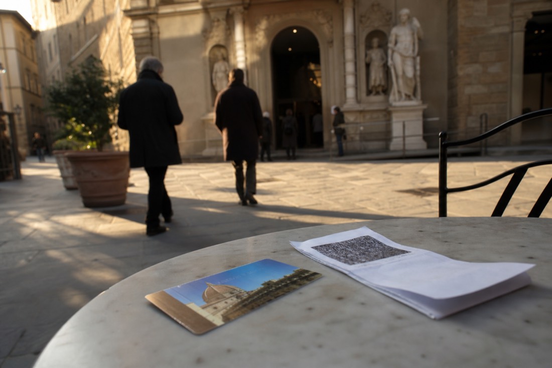 A Florence museum pass and a printed reservation slip resting on a cafe table with the Accademia entrance in the background