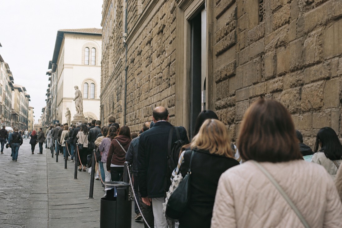 A long free-entry queue winding along Via Ricasoli past the Accademia facade on a bright Florence morning
