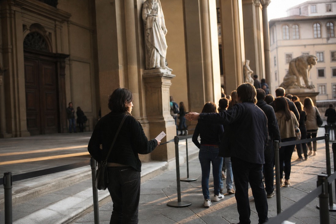 A puzzled visitor holding a museum pass at the entrance barrier of the Accademia while staff gesture toward the general line