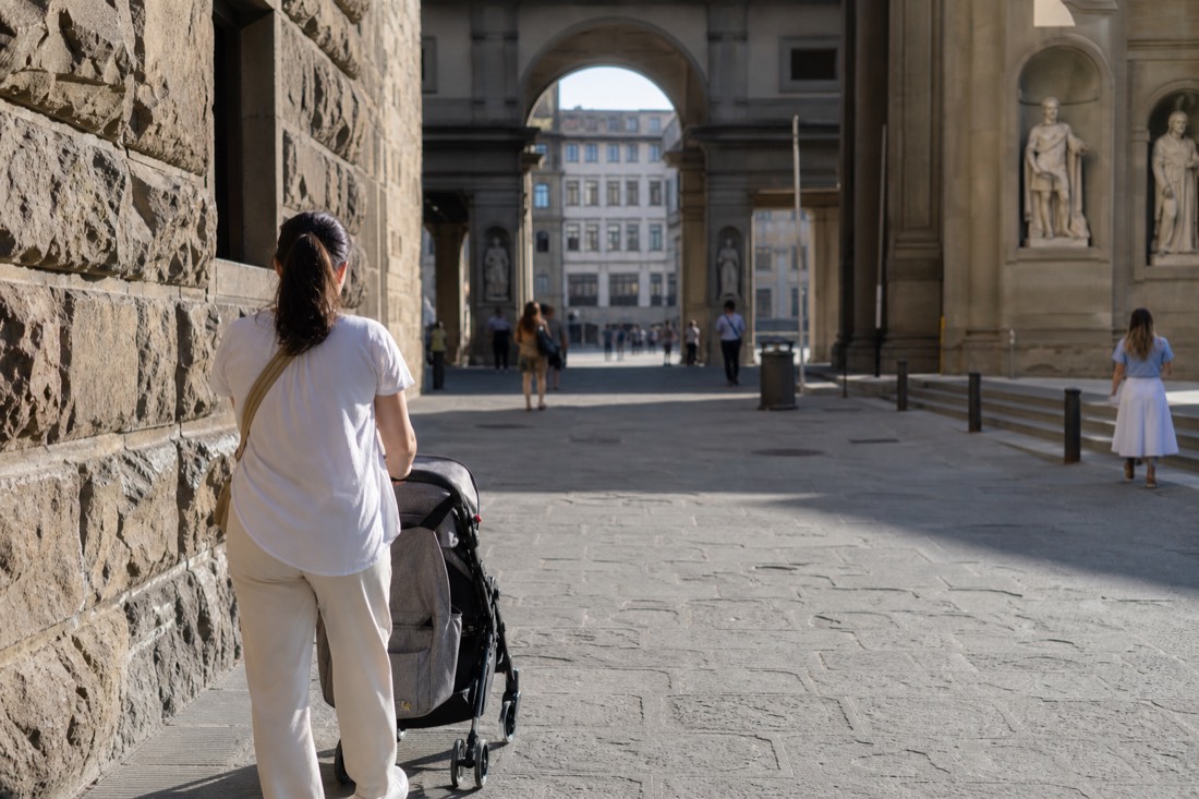 A parent pushing a stroller along a flat paved pedestrian street in central Florence toward the Accademia in soft morning light