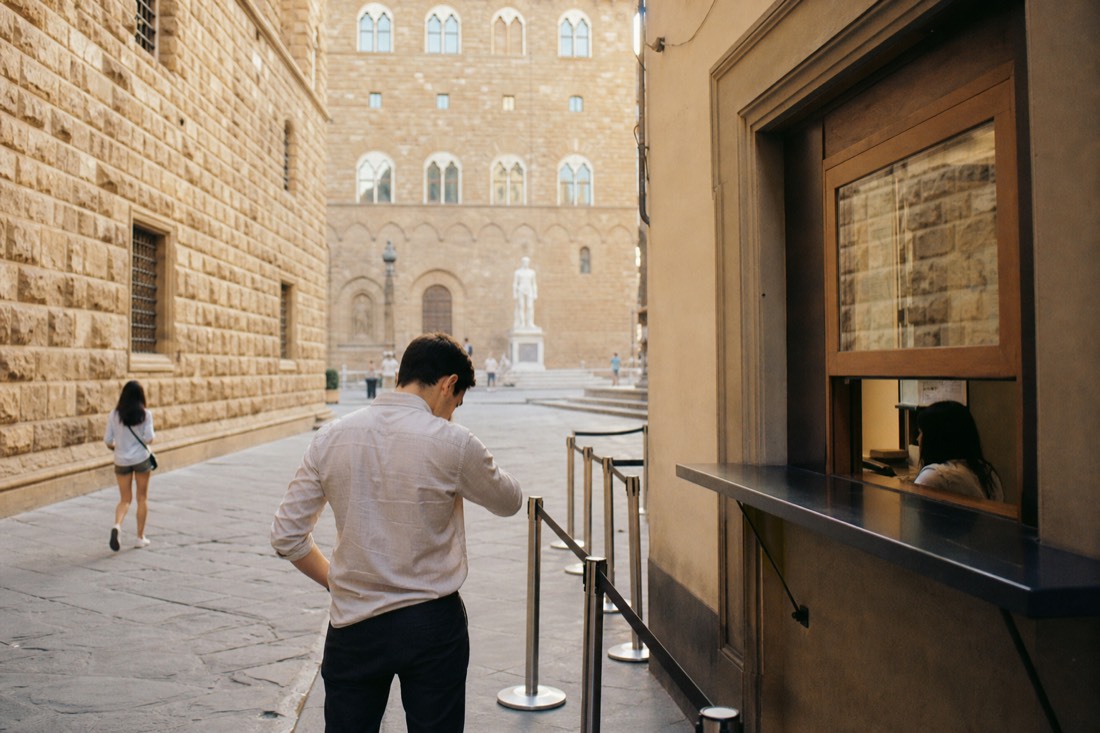 A visitor checking a wristwatch outside a ticket office on a quiet Florence street with the museum wall visible ahead