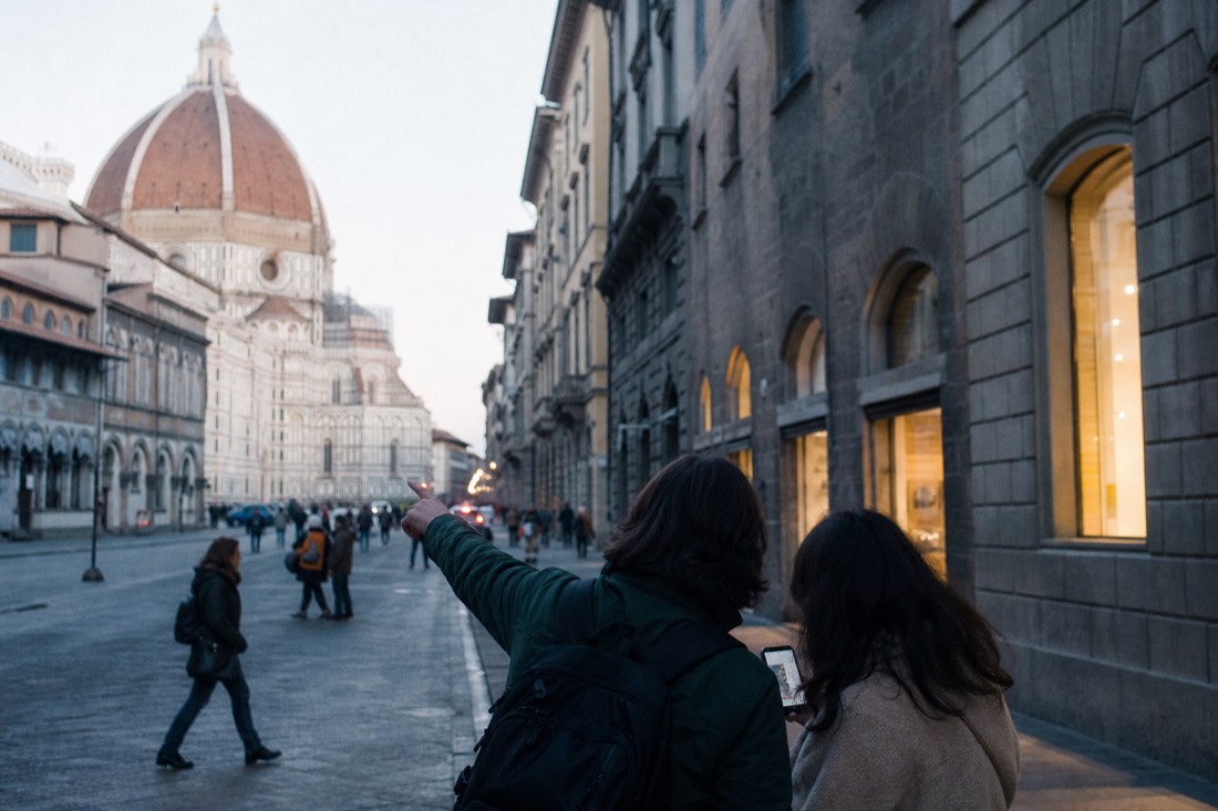 Two visitors comparing a phone map at a Florence street corner near the Duomo, one pointing up a long straight street