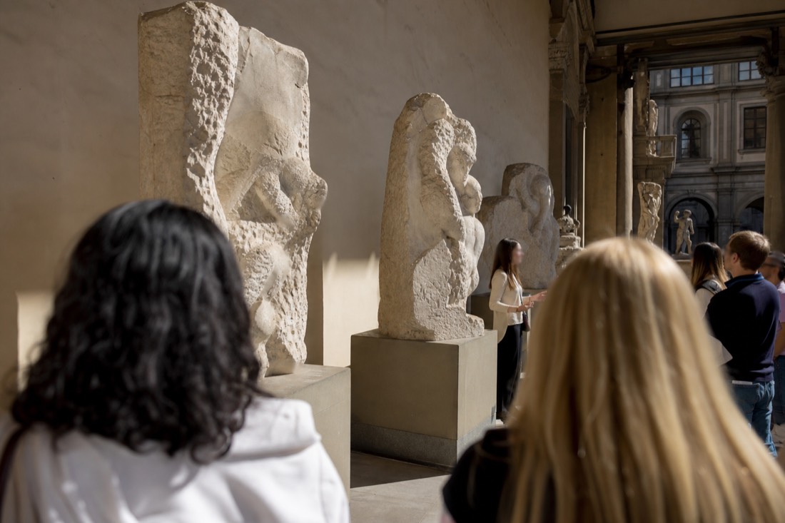 A small group gathered around a guide beside the unfinished Prisoners sculptures in the Accademia Gallery corridor in Florence