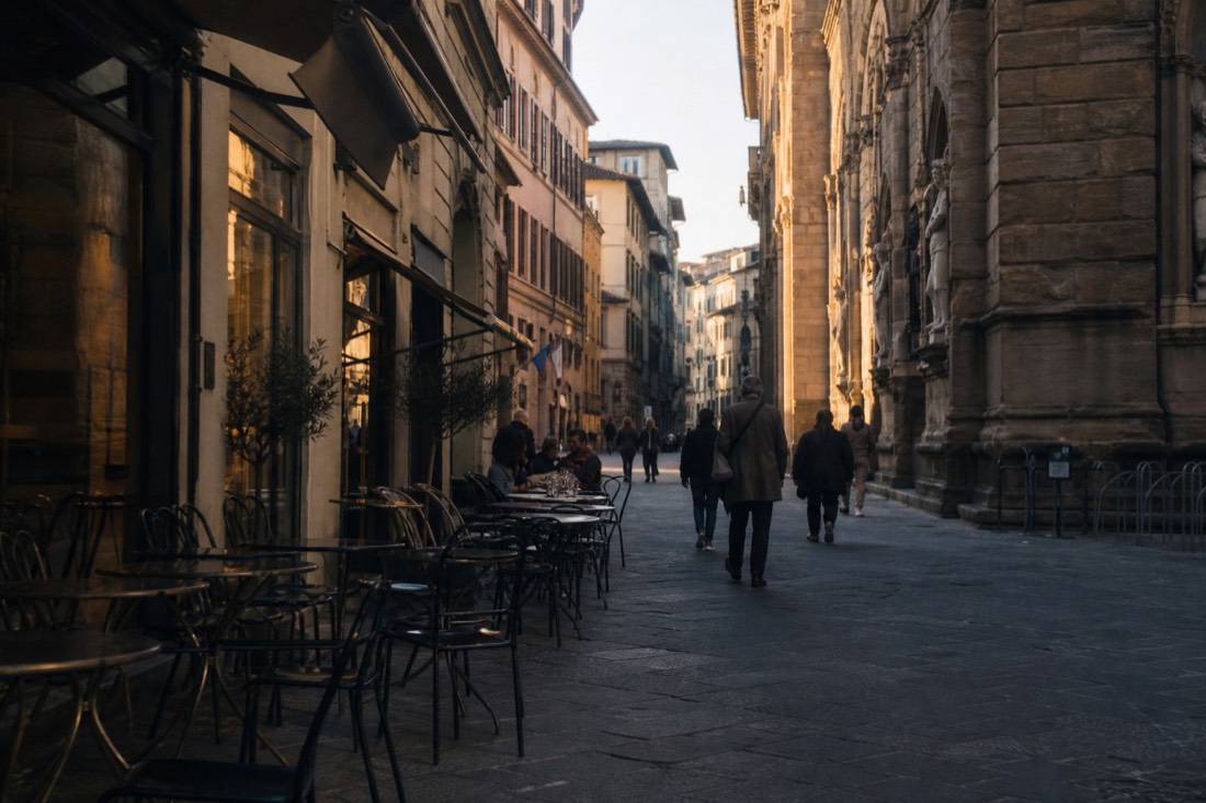 Late afternoon golden light on an emptying Florence street with cafe tables and the shadow of a museum facade