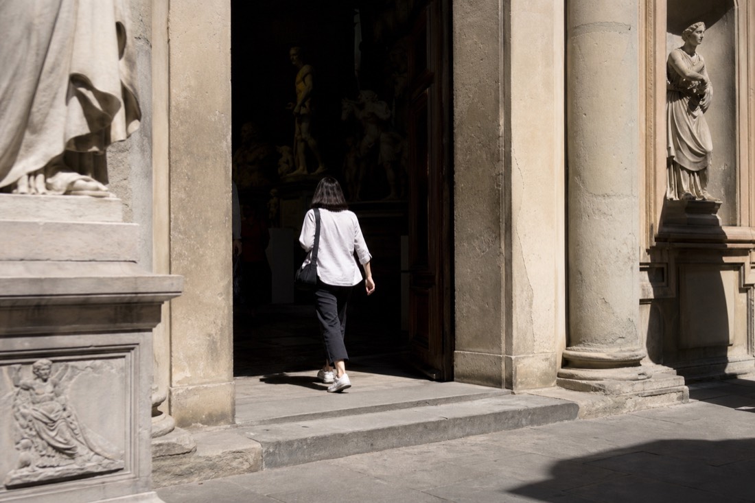 A visitor walking into the Accademia Gallery carrying only a small shoulder bag, hands otherwise free, in bright daylight