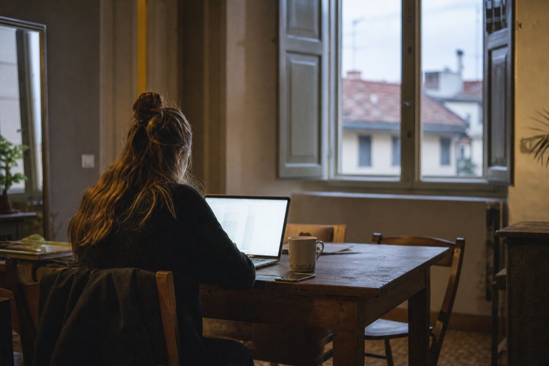 Glowing laptop screen on a rustic wooden table in a Florence apartment with shuttered windows at night