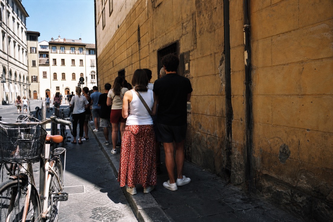 Short afternoon queue beside a weathered ochre wall on Via Ricasoli with bicycles parked along the curb