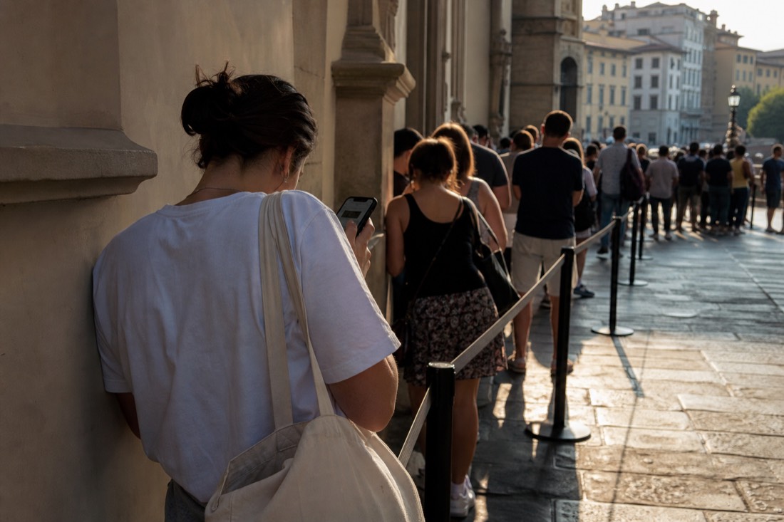 A tired traveler checking a phone at the back of a long museum queue in bright afternoon sun in Florence